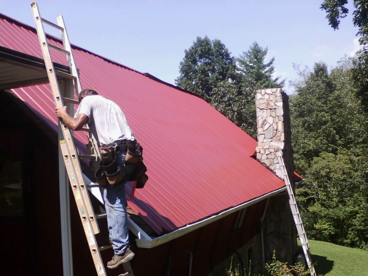 Roofer on a ladder beside a red metal roof and stone chimney — Soffit & Fascia Repair by Roofing Brothers in Malcolm